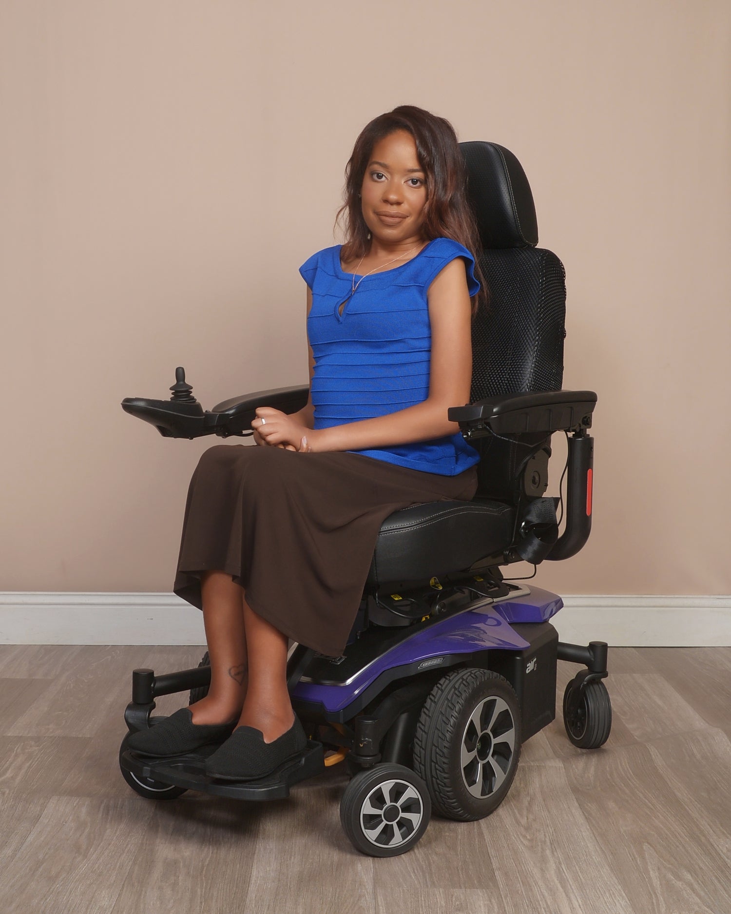 Woman in a blue top and brown skirt sitting in a black and purple power wheelchair against a beige wall.